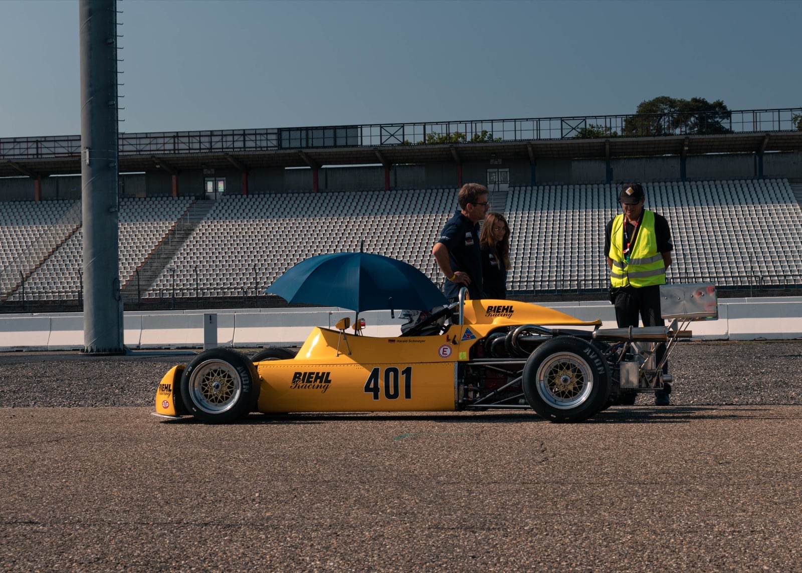 Gelber Vintage-Formel-Wagen Nummer 401 auf der Strecke vor leerer Tribüne, Personen mit Schirm im Briefing