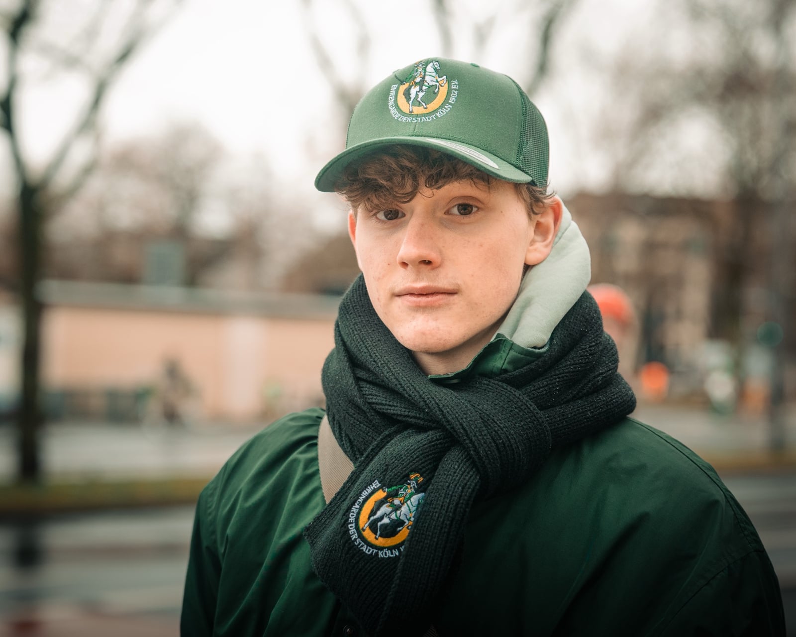 Portrait eines jungen Mannes mit grüner Schiffermütze und Schal der Brings im Stadtbild Kölns