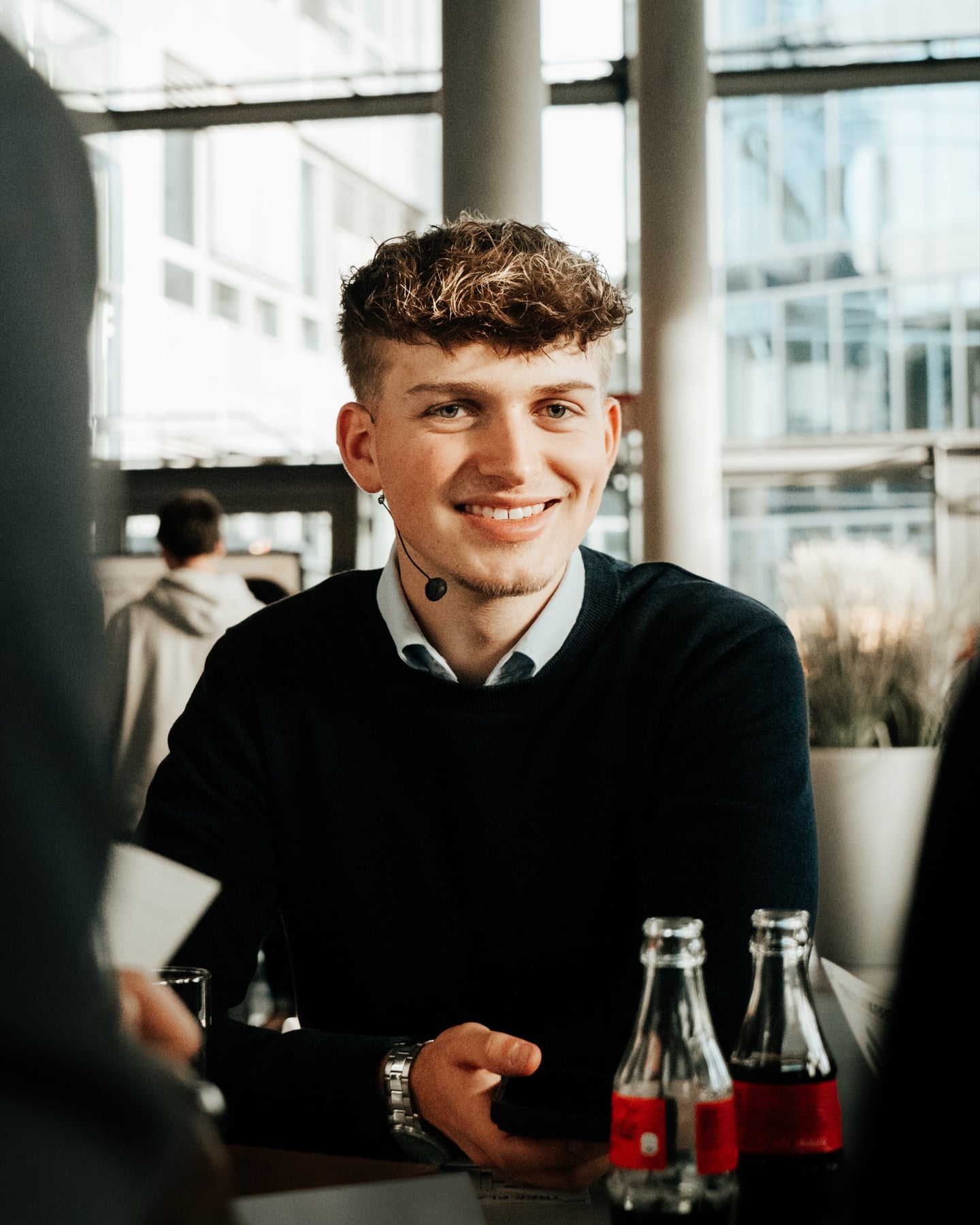 Junger Mann mit Locken und Headset im Gespräch beim SAP-Event, Coca-Cola-Flaschen im Vordergrund